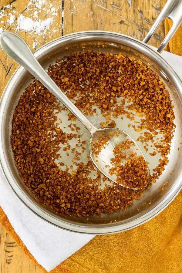 Toasted breadcrumbs in a saucepan with a spoon on a wooden surface.