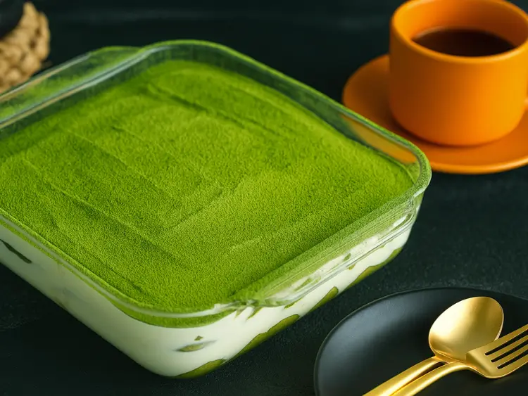 Chilled matcha tiramisu in a square glass dish with golden spoon and orange coffee cup