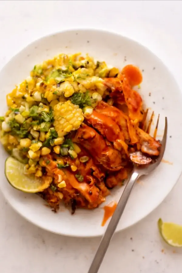 A plate of buffalo chicken served with a corn salad, garnished with lime and drizzled with sauce