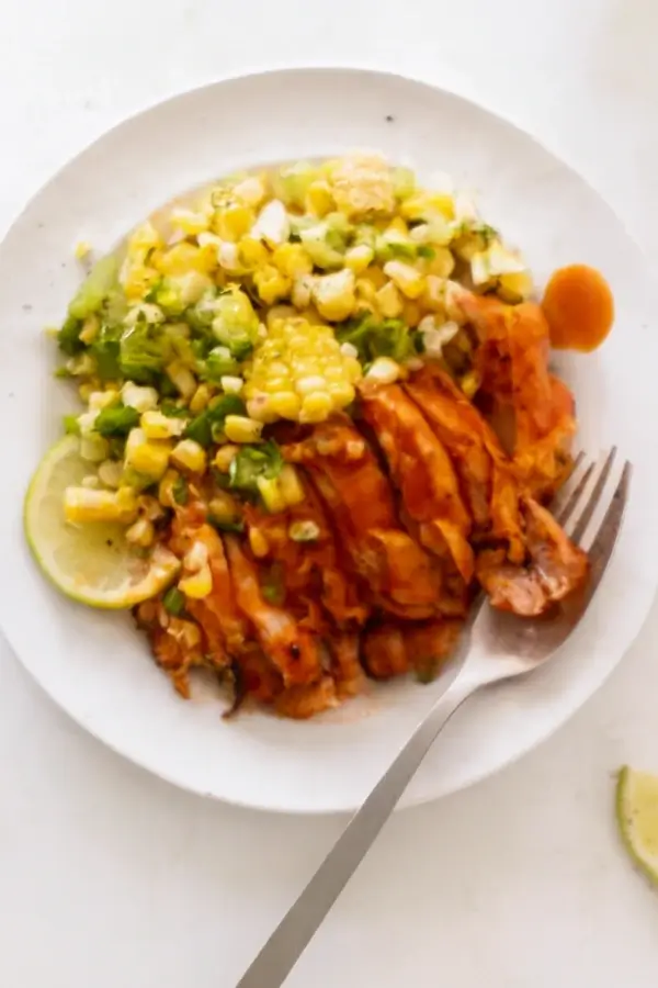 A plate with buffalo chicken thighs, corn salad, and lime wedges, with a fork in the foreground