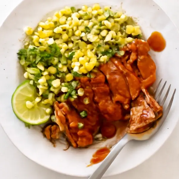 A plate with buffalo chicken, corn salad, and lime wedges with a fork on the side