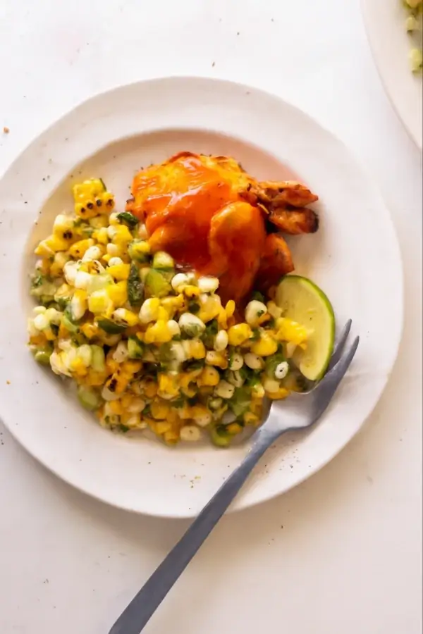 A close-up of buffalo chicken served with corn salad and a lime wedge on a plate
