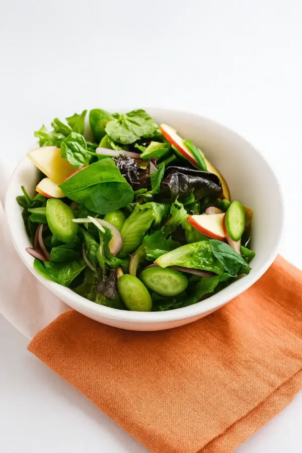 Korean Green Salad in a white bowl with mixed greens, cucumber rounds, red-skinned apple slices, and sesame seeds on an orange napkin