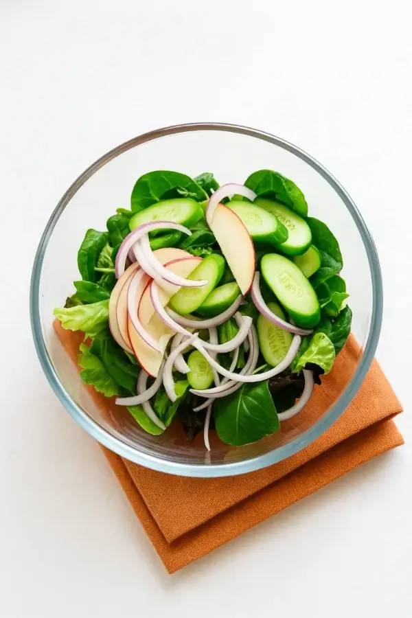Glass bowl filled with lettuce, cucumber slices, apple wedges, and red-onion strips before dressing